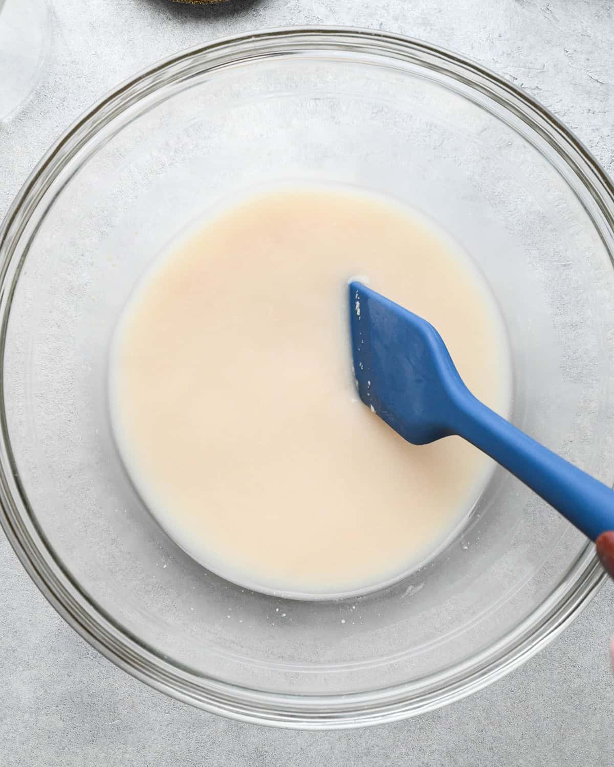 How to Make Pizza Dough - yeast, water and sugar in glass bowl on a gray surface with a blue spatula after mixing