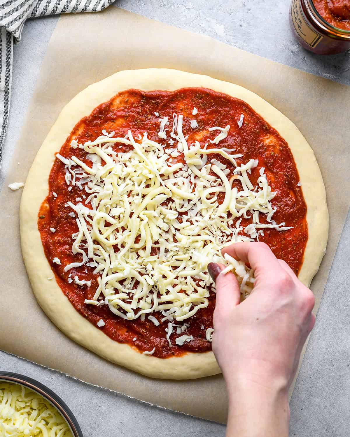 a hand sprinkling cheese over the pizza sauce on homemade pizza dough. The dough is on parchment paper and there is a bowl of cheese and jar of sauce nearby, with a striped towel in the top left corner.