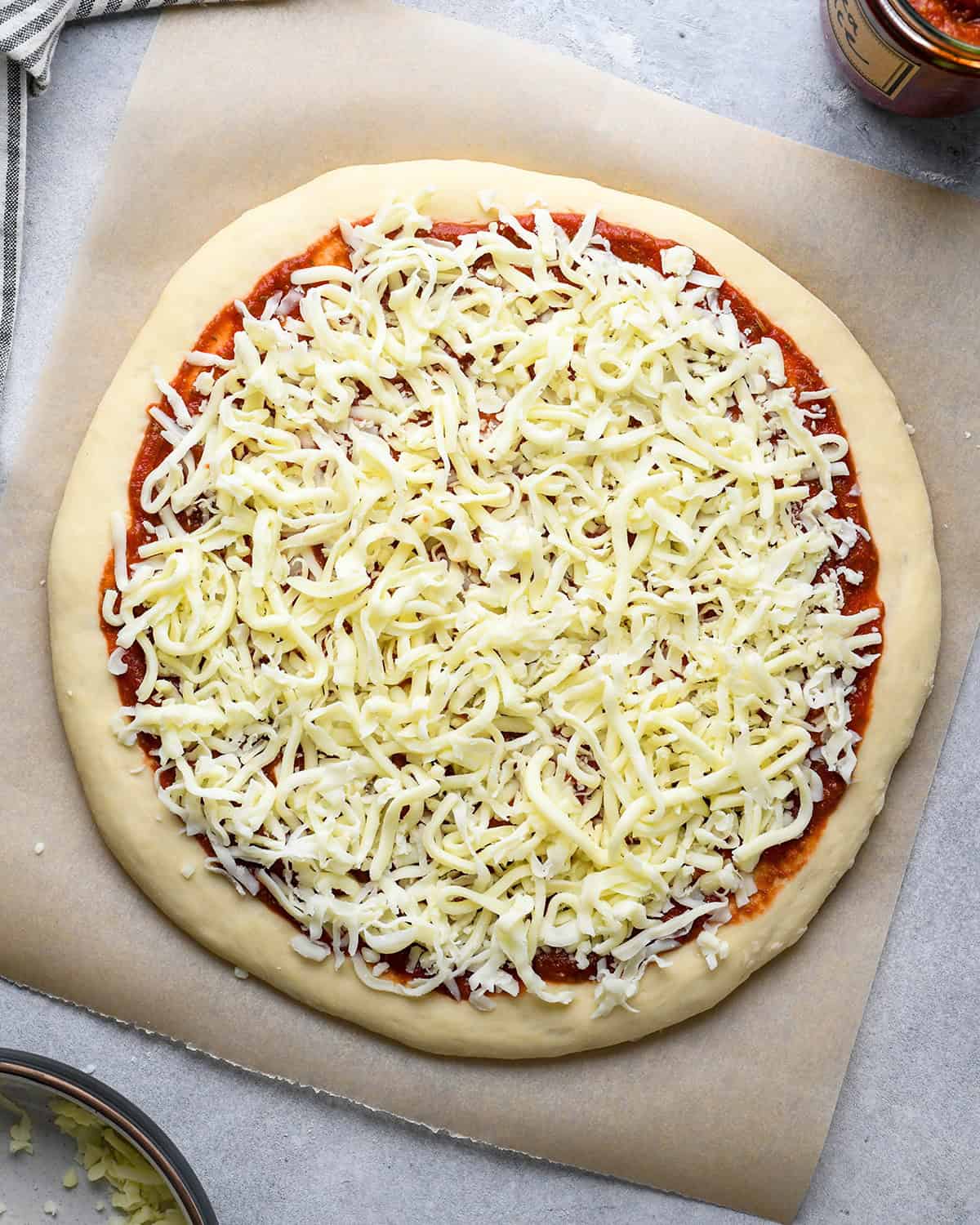 an assembled pizza using homemade pizza dough on parchment paper and there is a bowl of cheese and jar of sauce nearby, with a striped towel in the top left corner.