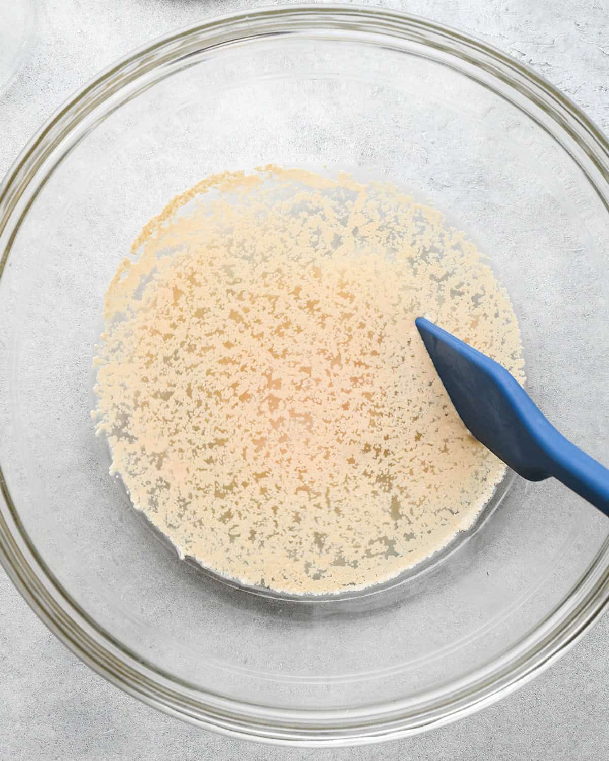 How to Make Pizza Dough - yeast, water and sugar in glass bowl on a gray surface with a blue spatula before mixing