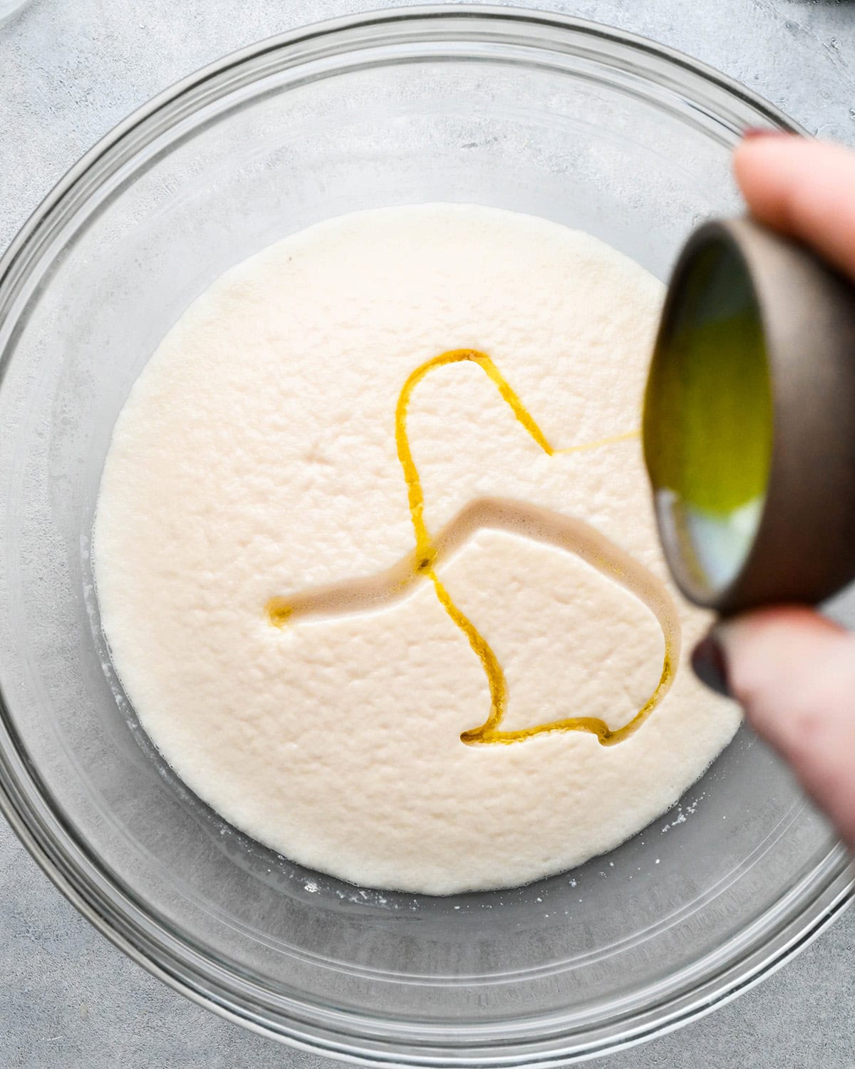 How to Make Pizza Dough - olive oil being added to the proofed yeast mixture in a glass bowl on a gray surface before mixing