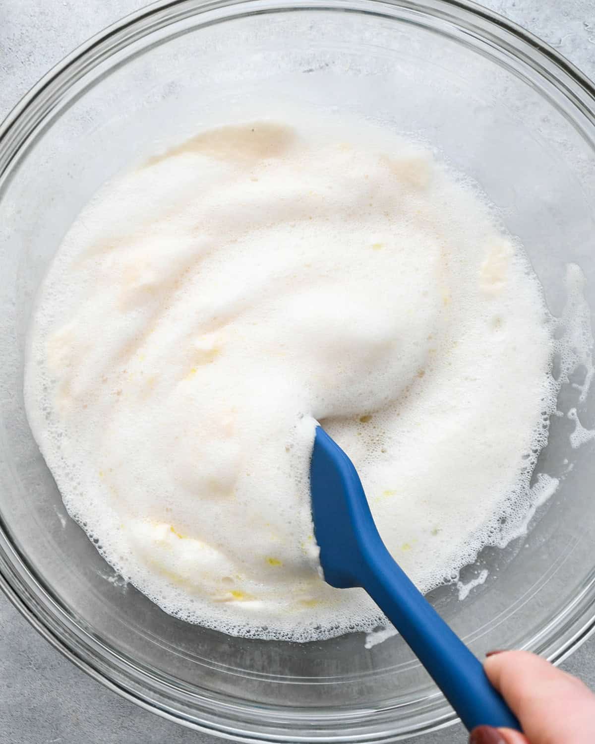 How to Make Pizza Dough - olive oil being mixed into the proofed yeast mixture in a glass bowl on a gray surface with a blue spatula