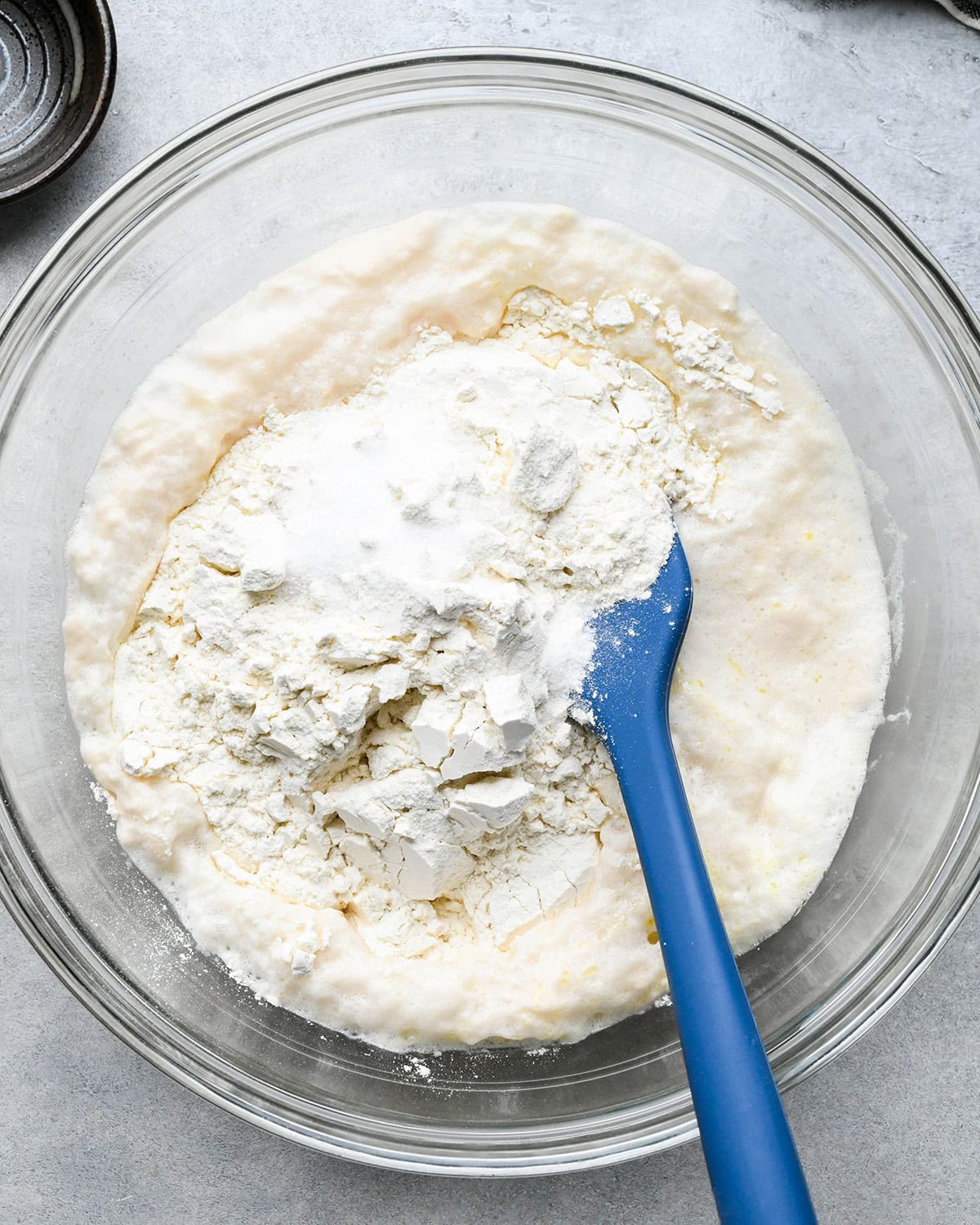 How to Make Pizza Dough - flour and salt added to the water and yeast mixture in a glass bowl on a gray surface with a blue spatula int he bowl