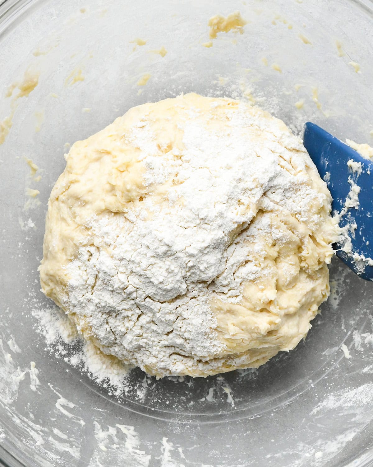 How to Make Pizza Dough flour added to the dough in a glass bowl with a blue spatula