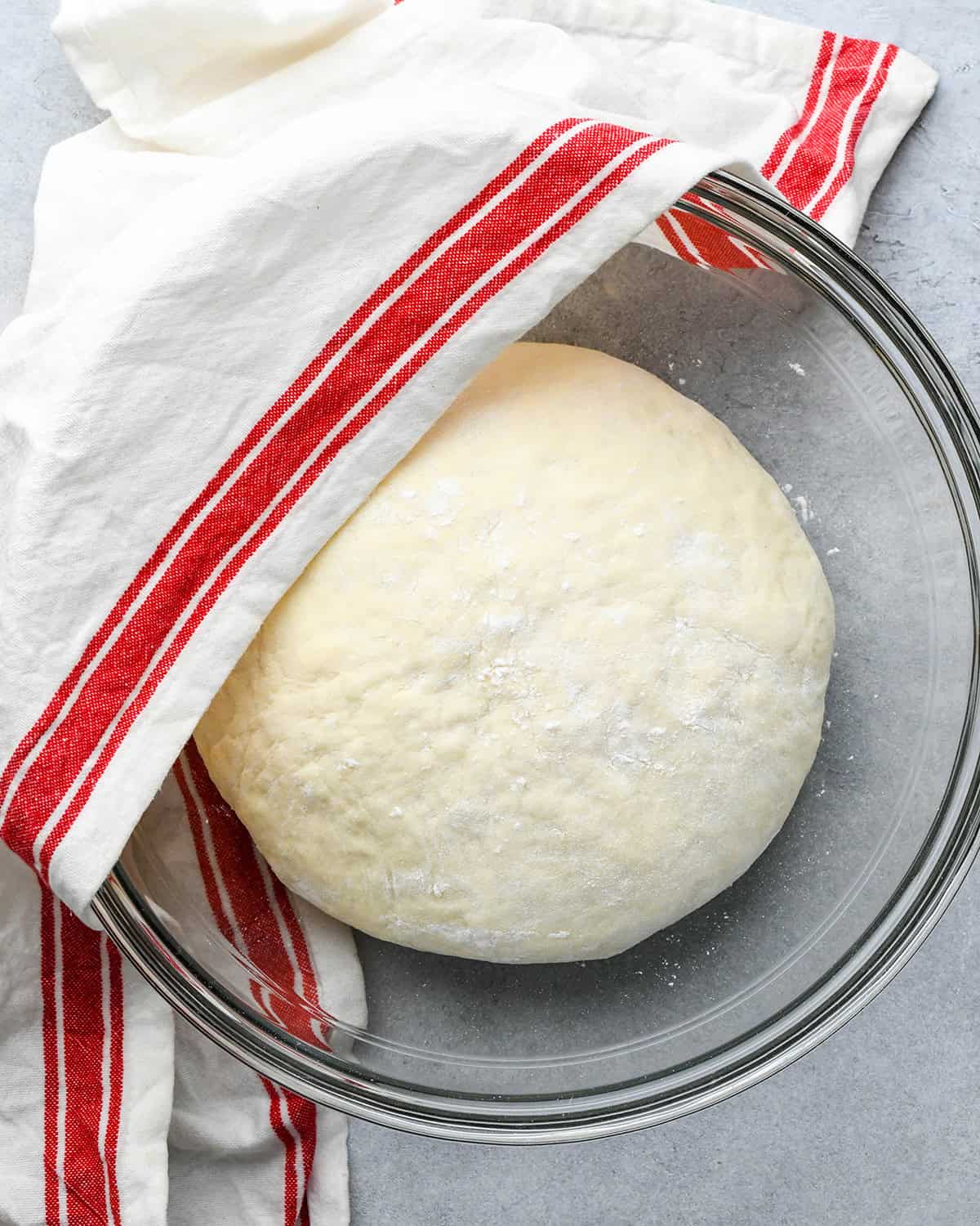 pizza dough in a bowl on a gray surface with a white and red tea towel partially covering the bowl and dough