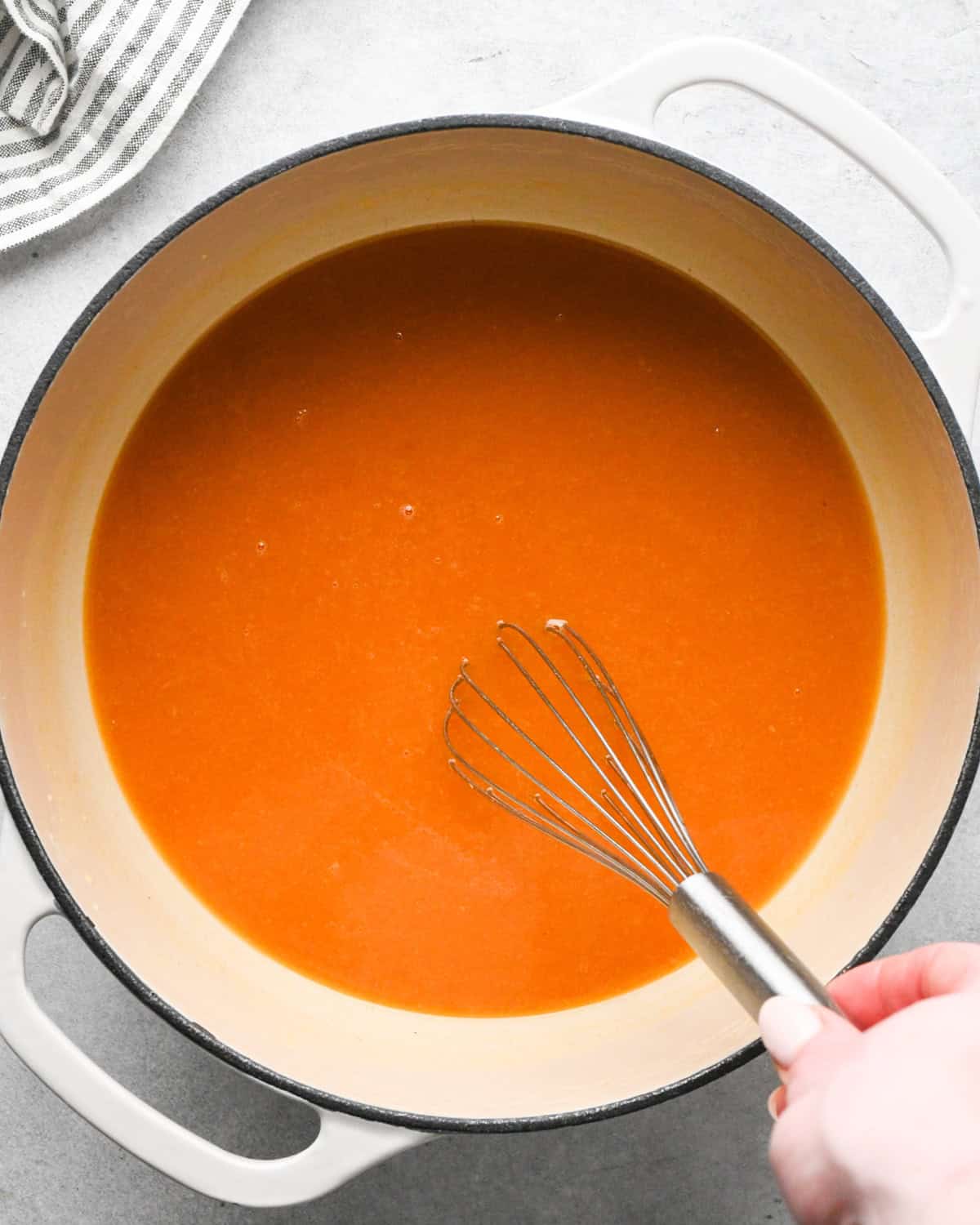 a hand whisking tomato soup in a white pot with a striped towel in the top left corner