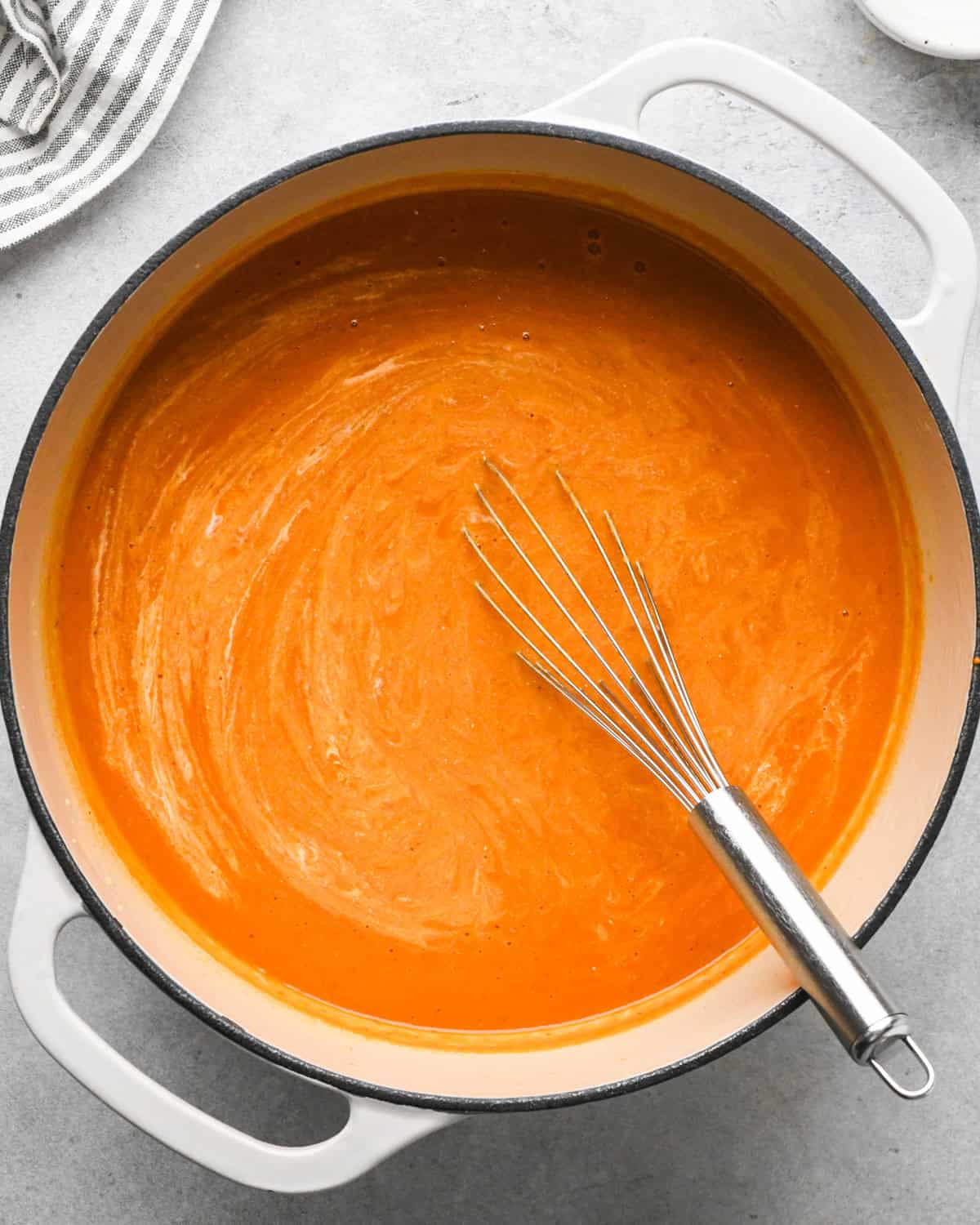 a pot of tomato soup with the heavy cream partially whisked into it, the whisk is in the pot and a striped towel sits beside the pot in the top left corner