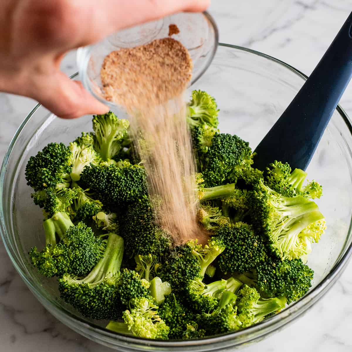 This is the best oven-roasted broccoli recipe! This easy roasted broccoli is made with 6 ingredients in 15 minutes! It's crispy, tender and perfectly seasoned! Baked broccoli is a healthy side dish that pairs well with everything! #broccoli #broccolirecipes #healthysidedish #sidedish #roastedbroccoli #joyfoodsunshine photo showing how to roast broccoli - pouring spice mixture onto oiled broccoli