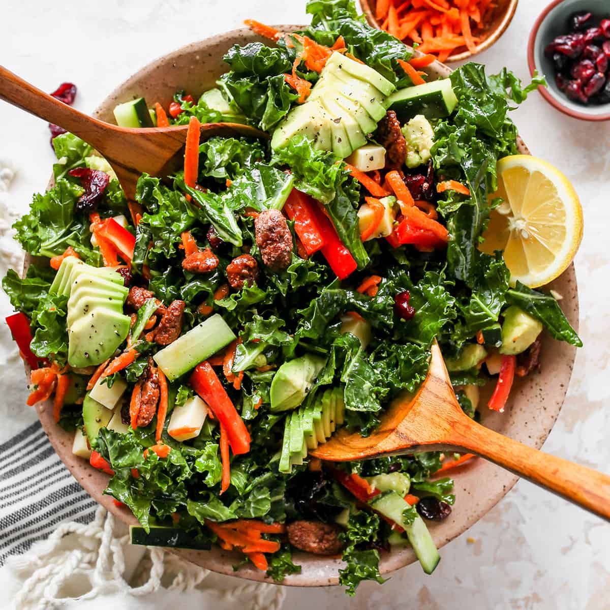 This kale salad recipe is loaded with colorful vegetables, sweet cranberries, crunchy pecans, creamy cheddar cheese and the best homemade dressing ever! It’s a hearty salad that holds up well and will impress even kale skeptics. overhead photo of wood spoons serving Kale Salad
