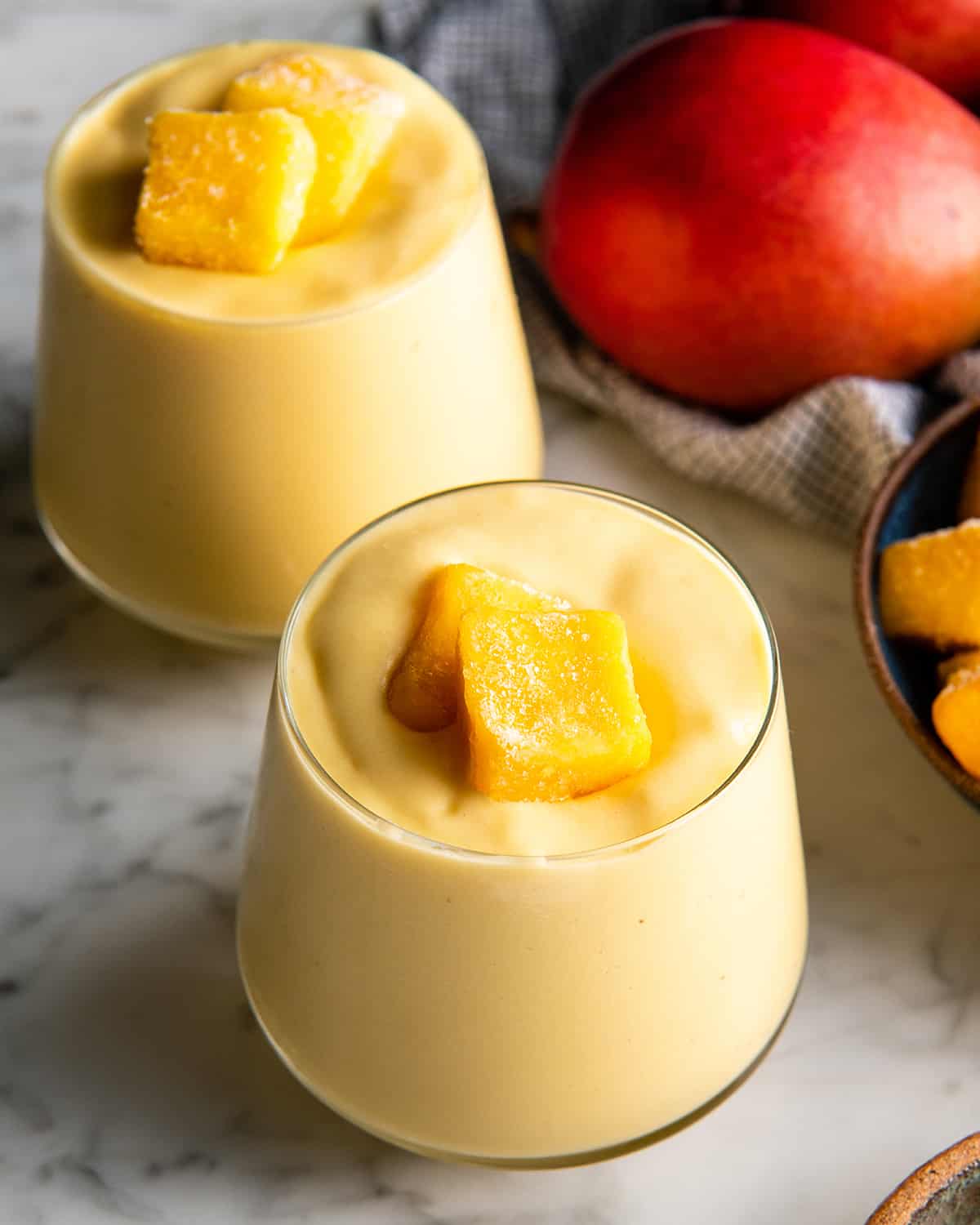The BEST mango smoothie recipe! This healthy & easy mango smoothie is made in 5 minutes with 6 ingredients (including coconut milk, yogurt and banana)! It's ultra creamy and you can use fresh or frozen mango and easily make it dairy-free! #smoothie #mango #mangosmoothie #healthy #recipe #joyfoodsunshine two glasses of mango smoothie with mango chunks on top