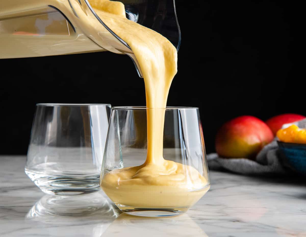 The BEST mango smoothie recipe! This healthy & easy mango smoothie is made in 5 minutes with 6 ingredients (including coconut milk, yogurt and banana)! It's ultra creamy and you can use fresh or frozen mango and easily make it dairy-free! #smoothie #mango #mangosmoothie #healthy #recipe #joyfoodsunshine front view of mango smoothie being poured into a glass from the blending container
