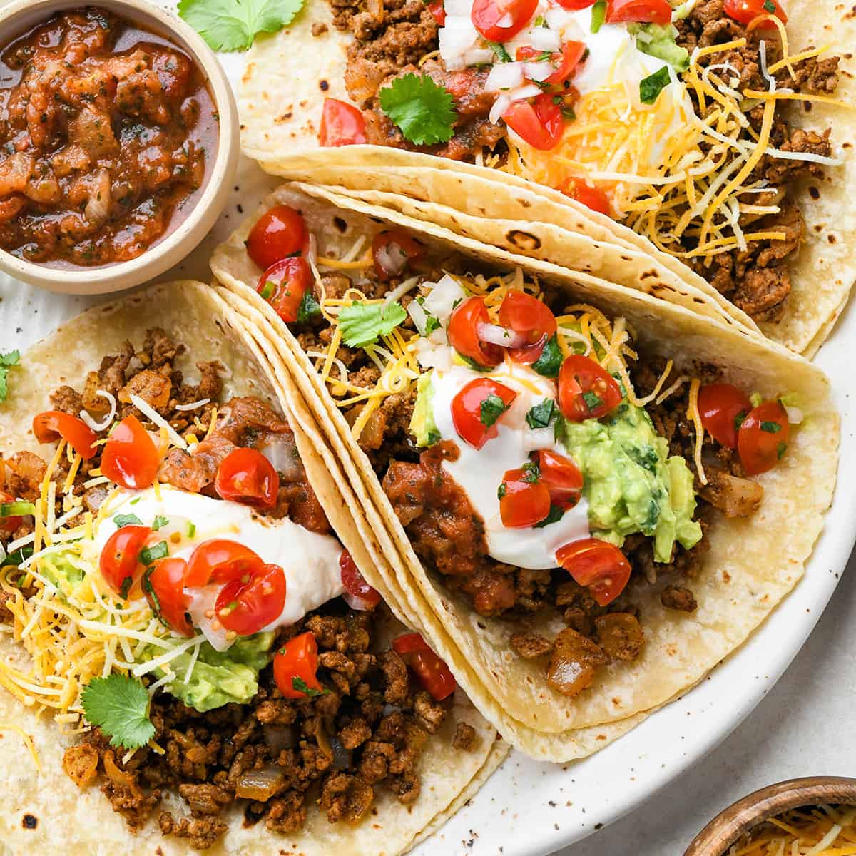 3 Mexican Ground Beef Tacos on a plate with sour cream, guacamole, cheese and tomatoes
