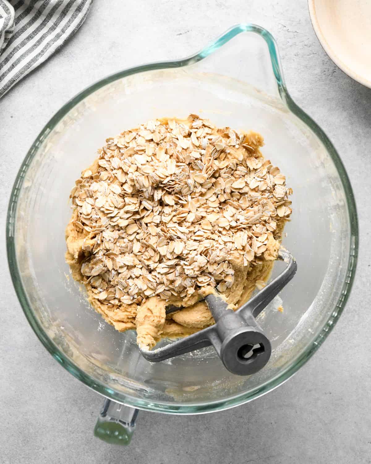 making chocolate chip oatmeal cookies - old fashioned oatmeal added to the dough before mixing in a glass bowl with a paddle attachement and a striped towel and bowl nearby on a gray surface