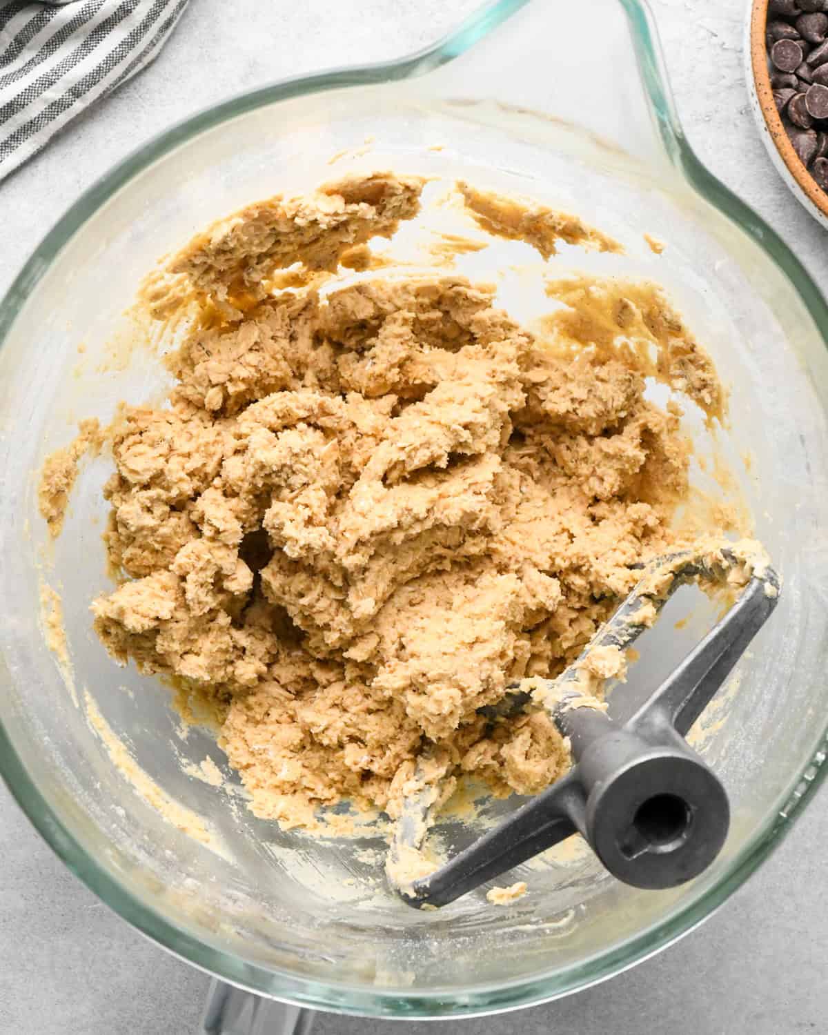 making chocolate chip oatmeal cookies - dough with oatmeal mixed in in a glass bowl with a paddle attachement and a striped towel and bowl of chocolate chips nearby