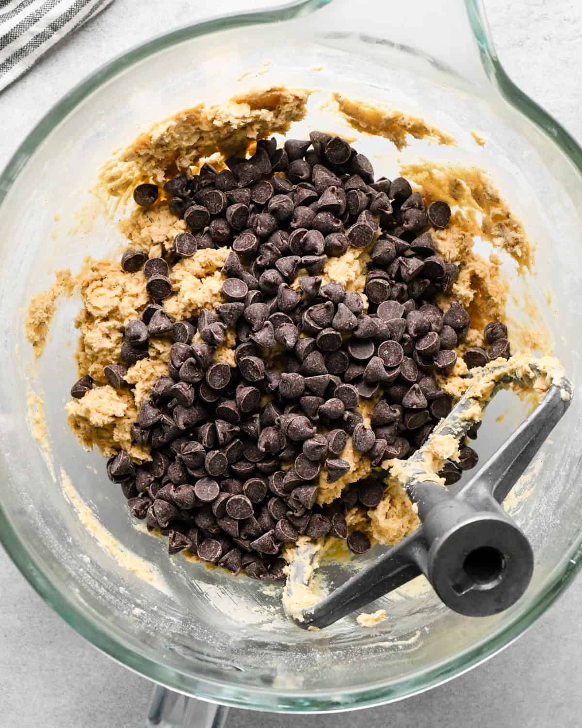 chocolate chips added to the cookie dough before mixing in a glass bowl with the paddle attachement on a gray surface with a striped towel nearby