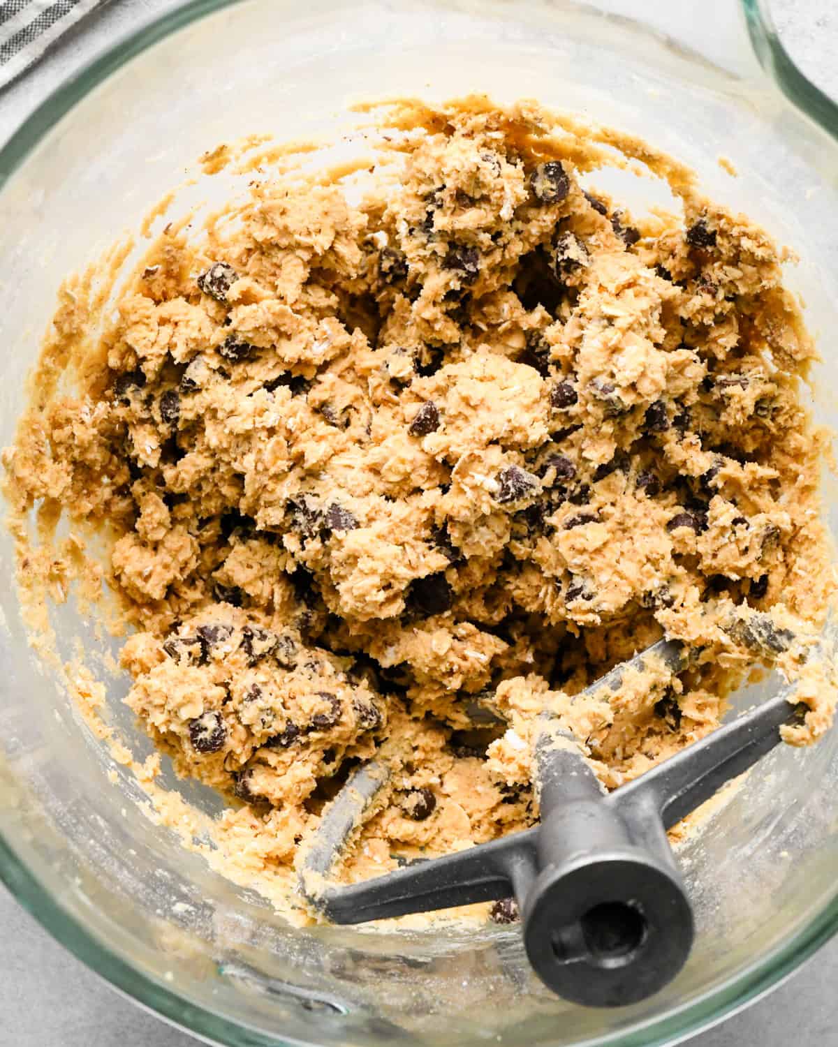 chocolate chip oatmeal cookie dough in a glass bowl with a paddle attachement and a striped towel in the top left corner.