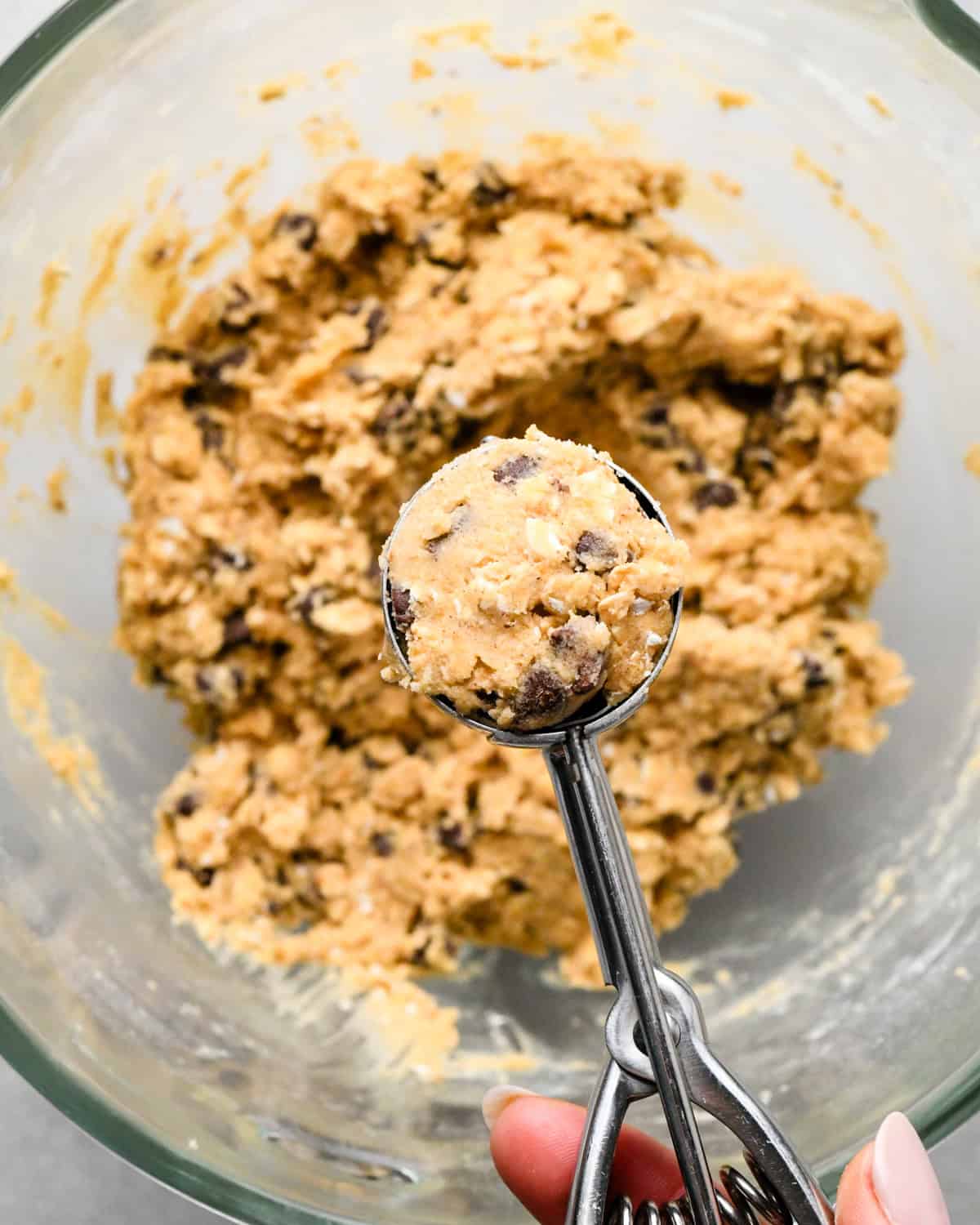 a hand holding a cookie scoop with a 2 Tablespoon portion of oatmeal chocolate chip cookie dough being held over a bowl of dough