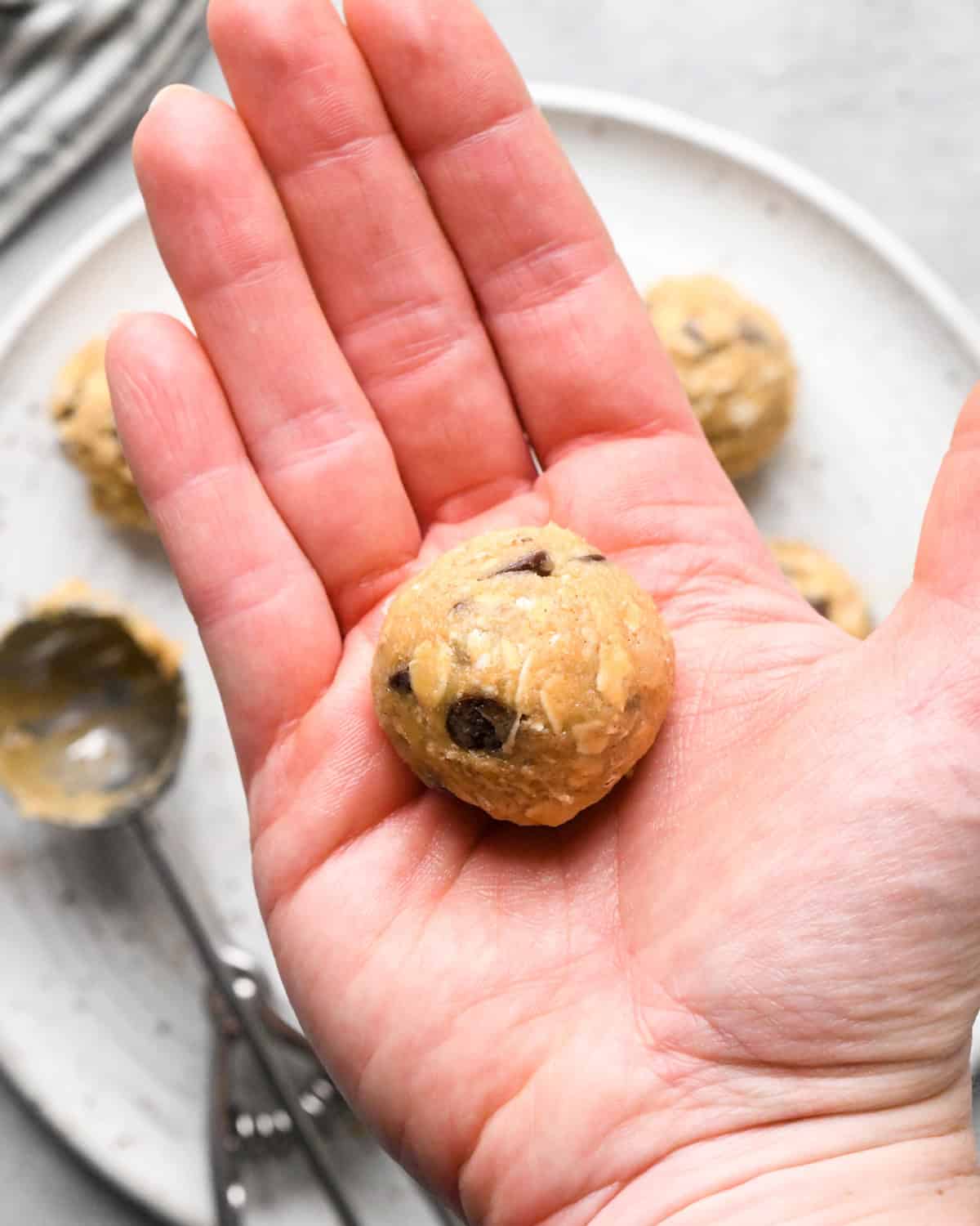 a ball of chocolate chip oatmeal cookie dough in a hand hovering over a plate of rolled cookies and an empty cookie scoop
