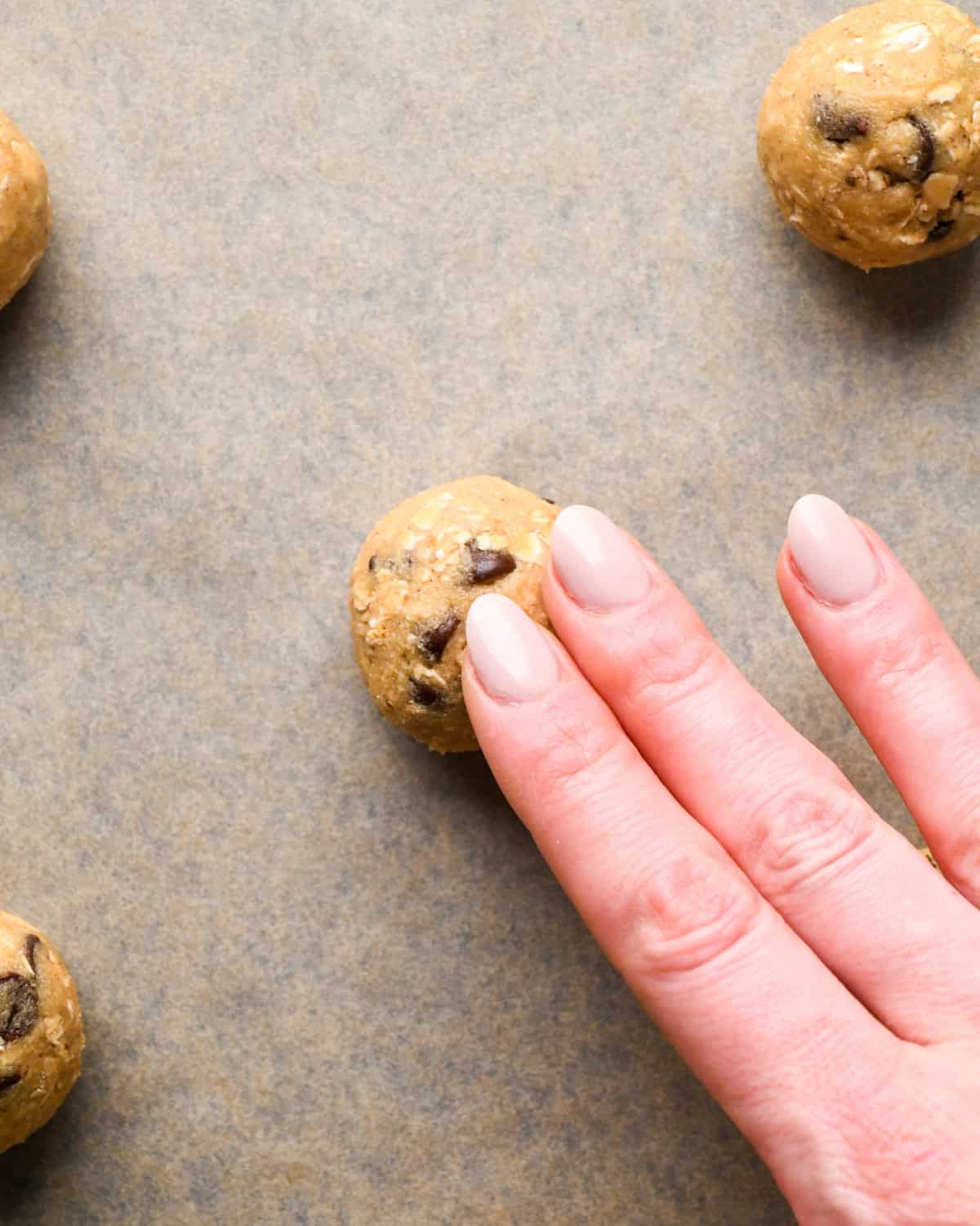 a hand pressing down the top of a chocolate chip oatmeal cookie dough ball on parchment paper on a baking sheet surrounded by 3 other dough balls