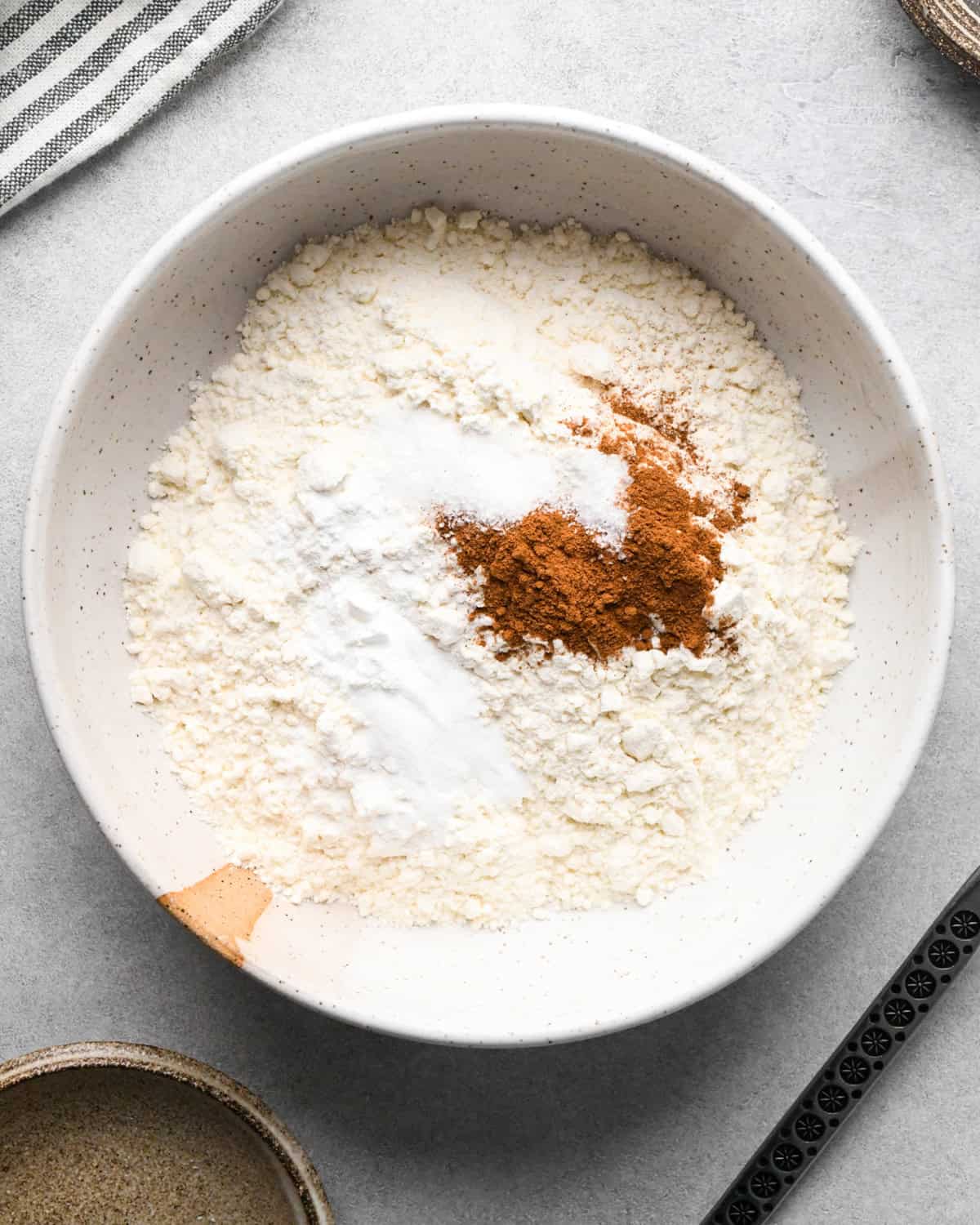 a bowl of flour, salt, baking soda, baking powder and cinnamon before the ingredients are mixed on a gray surface with a striped towel, bowl and spoon nearby