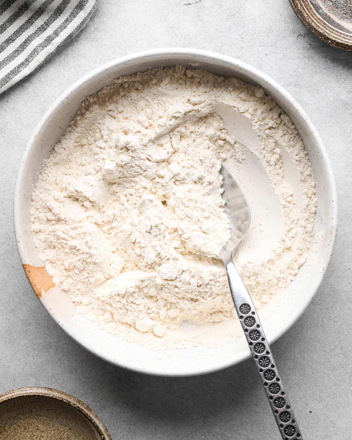 a bowl of flour, salt, baking soda, baking powder and cinnamon after the ingredients are mixed on a gray surface with a spoon int he flour stirring