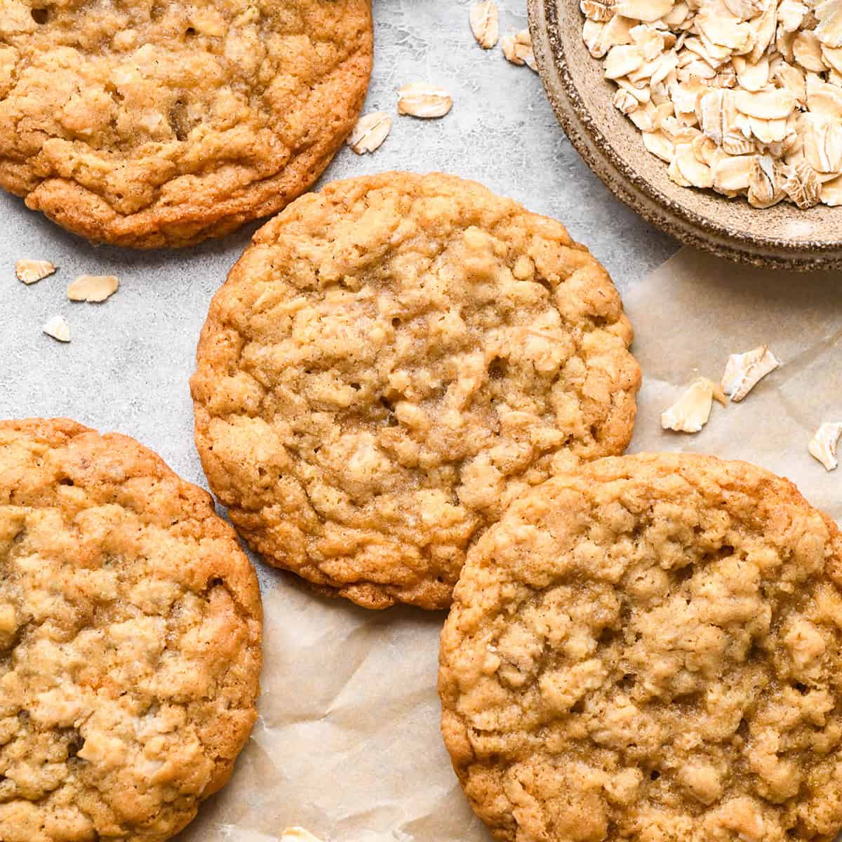 The BEST Oatmeal Cookie Recipe - crispy edges with soft and chewy centers, these oatmeal cookies are easy to make and out-of-this-world delicious. Add your favorite mix-ins for an extra special twist! #oatmealcookies #cookies #baking four oatmeal cookies on parchment paper next to a bowl of raw oats
