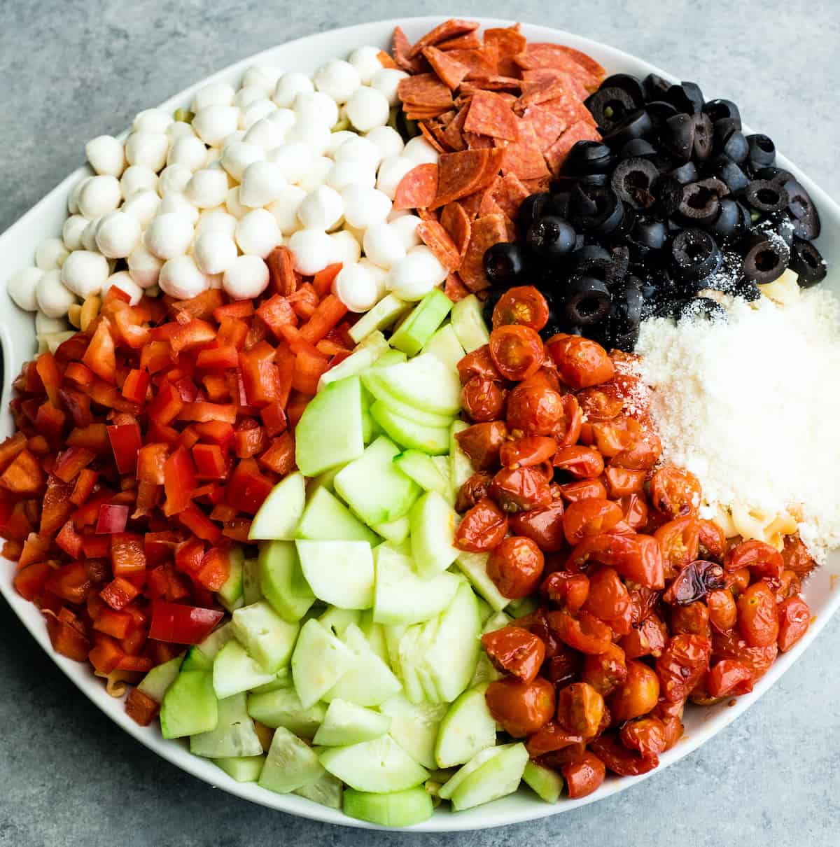 This Best Pasta Salad Recipe is made with Homemade Dressing & Roasted Tomatoes! It is the perfect healthy & flavorful make-ahead BBQ side dish full of fresh veggies, pepperoni and fresh mozzarella cheese! #pastasalad #bbq #sidedish #bestpastasalad #pastasaladrecipe #recipe #healthyrecipe #easypastasalad #homemadedressing overhead view of the best pasta salad recipe in a large white serving bowl with all the ingredients organized separately before being mixed together