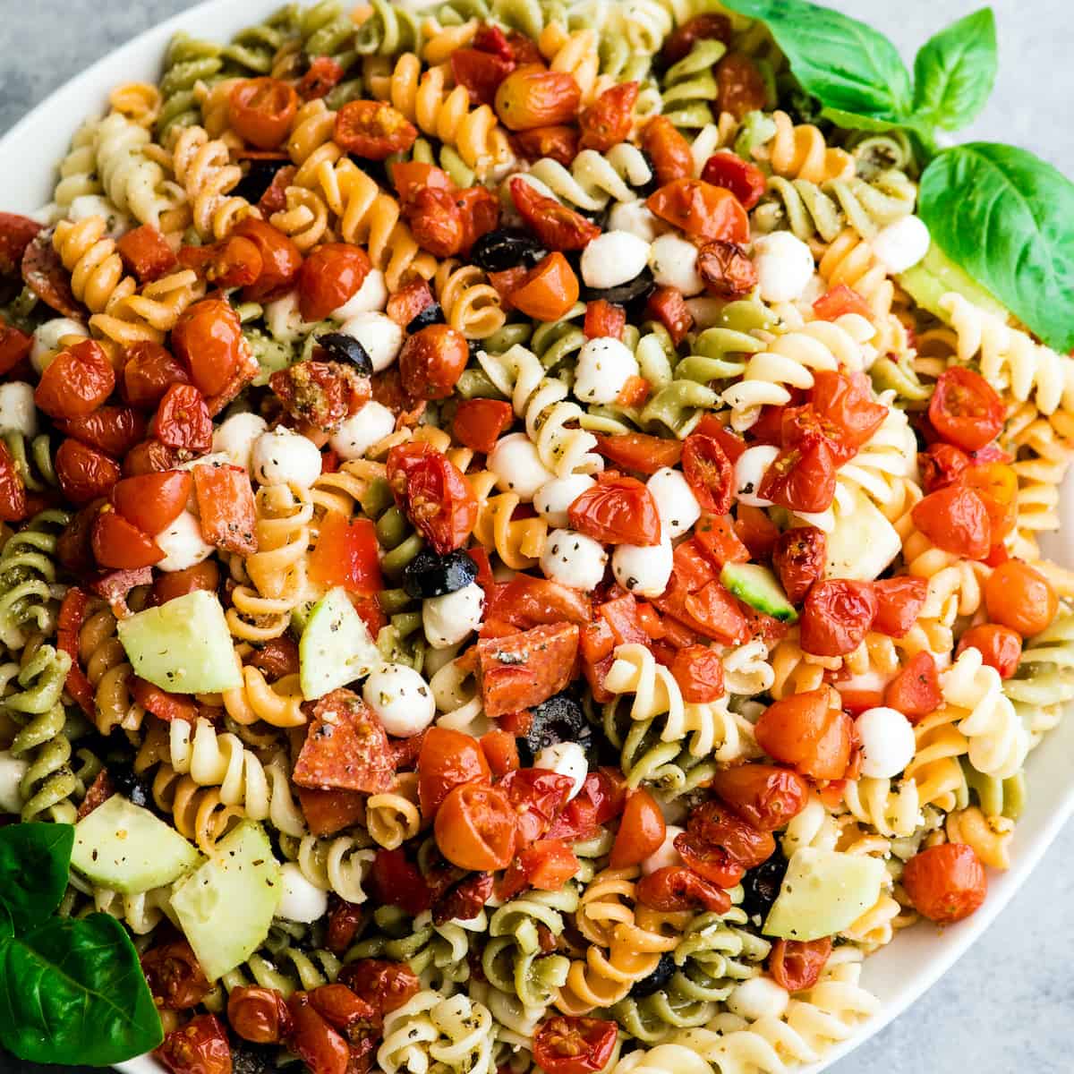 This Best Pasta Salad Recipe is made with Homemade Dressing & Roasted Tomatoes! It is the perfect healthy & flavorful make-ahead BBQ side dish full of fresh veggies, pepperoni and fresh mozzarella cheese! #pastasalad #bbq #sidedish #bestpastasalad #pastasaladrecipe #recipe #healthyrecipe #easypastasalad #homemadedressing overhead view of the best pasta salad recipe in a large white serving bowl with a wooden spoon taking a scoop