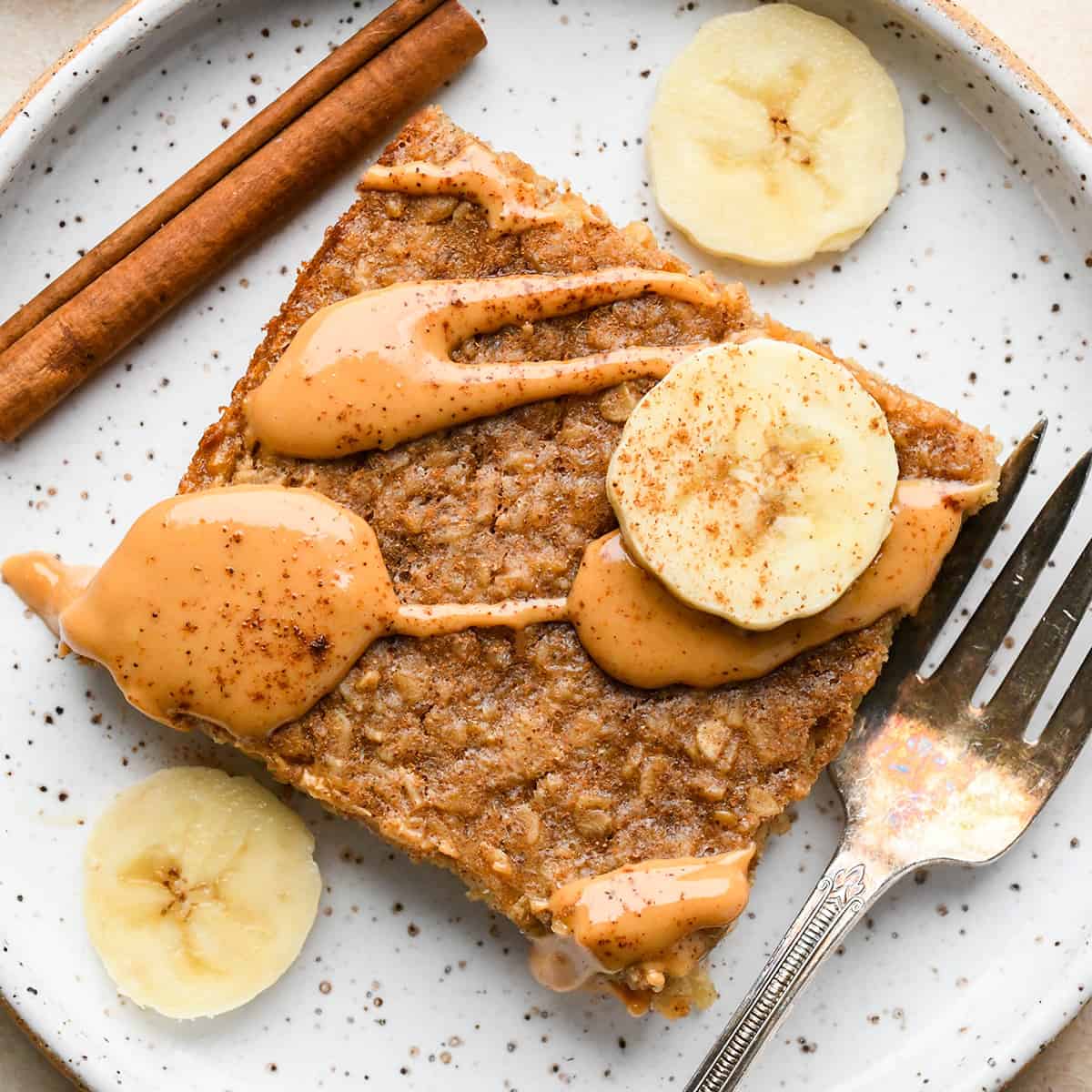 This Healthy Peanut Butter Banana Baked Oatmeal is the perfect make-ahead breakfast recipe! It's gluten-free, dairy-free, & vegan-friendly with no refined sugar! #peanutbutter #bakedoatmeal #banana #breakfast #recipe #glutenfree #dairyfree #veganfriendly #kidfriendly #healthyrecipe a piece of Peanut Butter Banana Baked Oatmeal on a plate with a drizzle of peanut butter, bananas and a fork