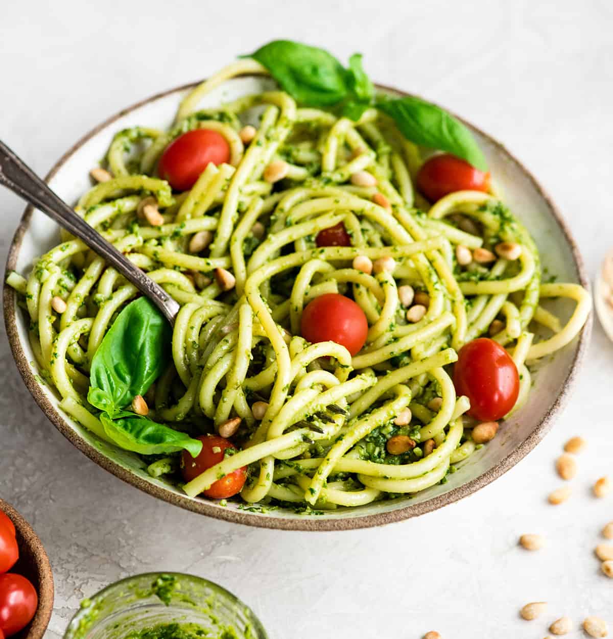 This pesto pasta recipe is a delicious quick and easy summer recipe! It can be served as a main dish or side dish, and you can enjoy it warm or cold! Full instructions how to make your own basil pesto are included, or you can make it even easier and use store-bought! #pestopasta #pesto #basil #summerrecipe #summer #toastedpinenuts #pinenuts #pasta Overhead/front view of a bowl of Pesto Pasta with tomatoes, fresh basil and toasted pine nuts and a fork with a twirl of pasta on it