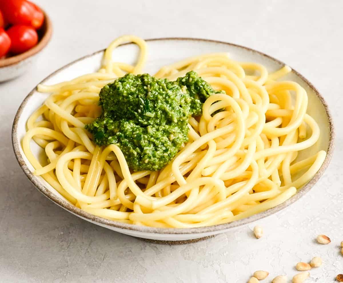 This pesto pasta recipe is a delicious quick and easy summer recipe! It can be served as a main dish or side dish, and you can enjoy it warm or cold! Full instructions how to make your own basil pesto are included, or you can make it even easier and use store-bought! #pestopasta #pesto #basil #summerrecipe #summer #toastedpinenuts #pinenuts #pasta front view of a homemade pesto sauce on bowl of spaghetti noodles to make Pesto Pasta before mixing in