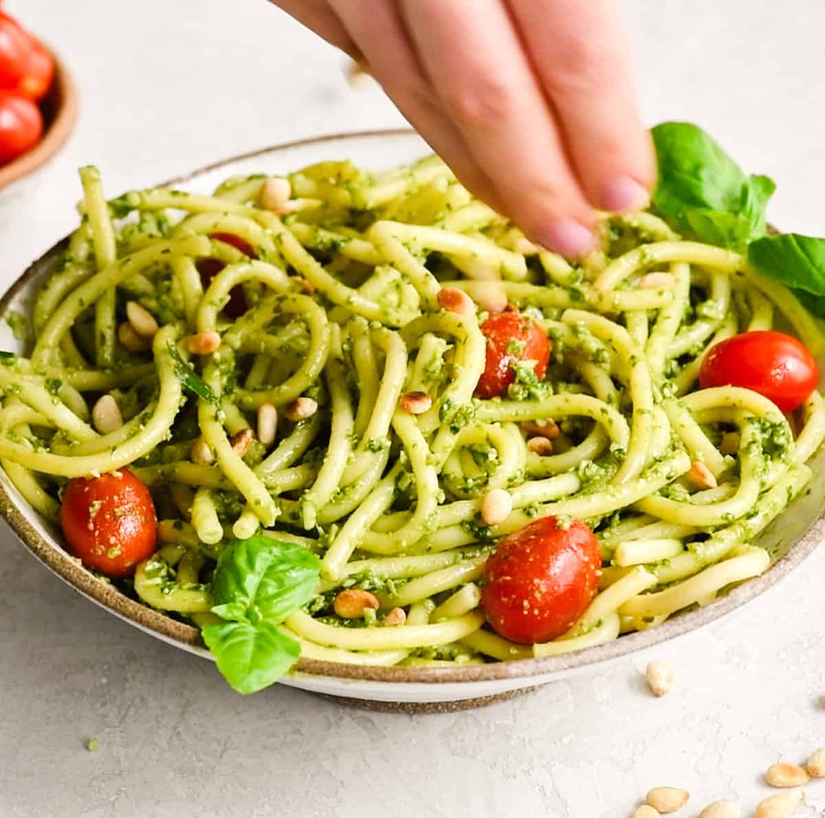 This pesto pasta recipe is a delicious quick and easy summer recipe! It can be served as a main dish or side dish, and you can enjoy it warm or cold! Full instructions how to make your own basil pesto are included, or you can make it even easier and use store-bought! #pestopasta #pesto #basil #summerrecipe #summer #toastedpinenuts #pinenuts #pasta pine nuts being sprinkled over a bowl of pesto pasta