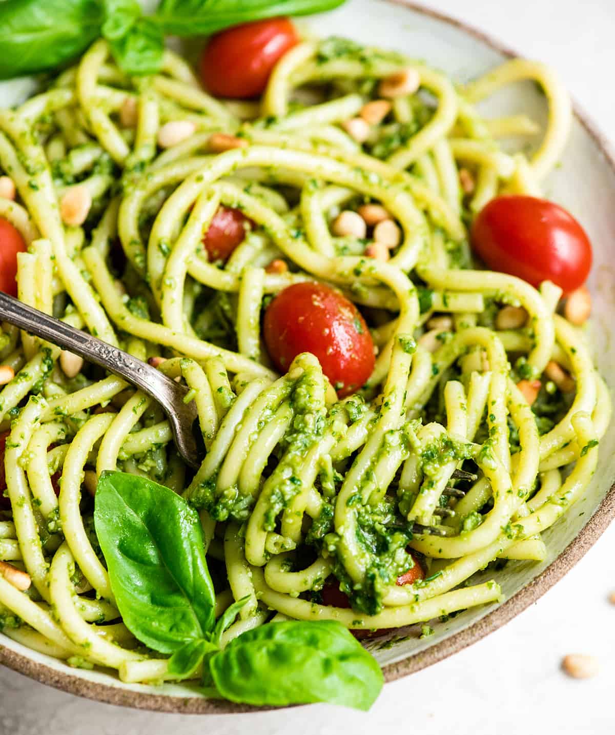 This pesto pasta recipe is a delicious quick and easy summer recipe! It can be served as a main dish or side dish, and you can enjoy it warm or cold! Full instructions how to make your own basil pesto are included, or you can make it even easier and use store-bought! #pestopasta #pesto #basil #summerrecipe #summer #toastedpinenuts #pinenuts #pasta front view of a bowl of Pesto Pasta with tomatoes, fresh basil and toasted pine nuts and a fork with a twirl of pasta on it