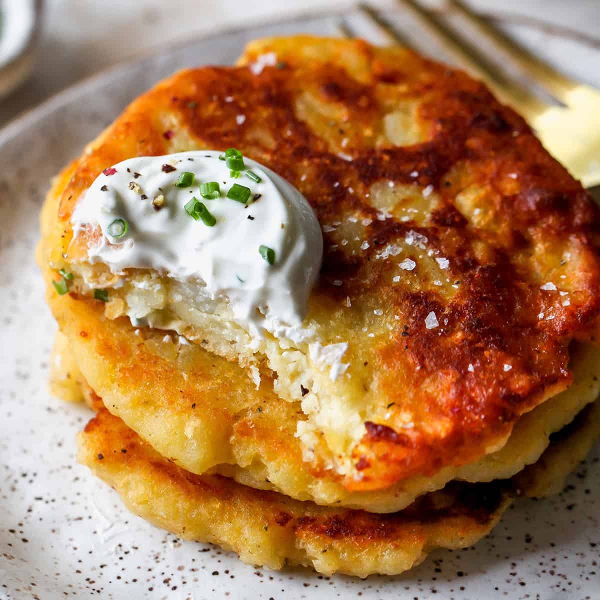 a stack of 3 Potato Pancakes, the top one has a bite taken out of it. Garnished with sour cream and chives.