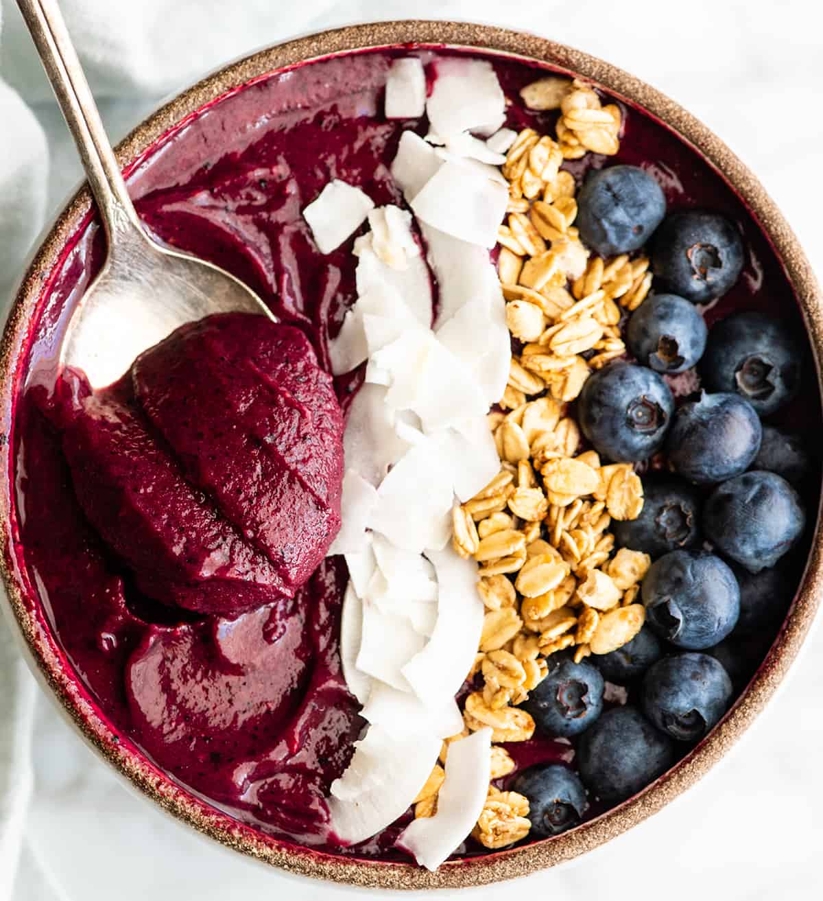 Follow our step-by-step photos & video to learn how to make the BEST easy & healthy smoothie bowl recipe (3 varieties) with 5 ingredients in 5 minutes! Smoothie bowls make the best breakfast, brunch or snack, and you can save so much money making them at home! Paleo, dairy-free and vegan-friendly. Overhead view of an acai smoothie bowl with toppings and a spoon taking a bite out of it