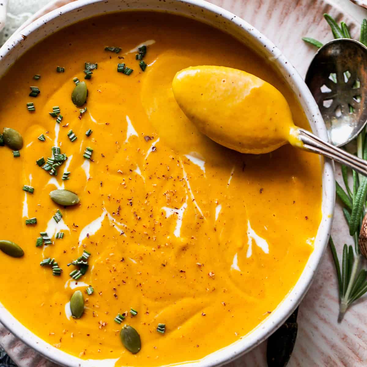 a spoon taking a bite out of a bowl of Sweet Potato Soup garnished with rosemary, cream and seeds.