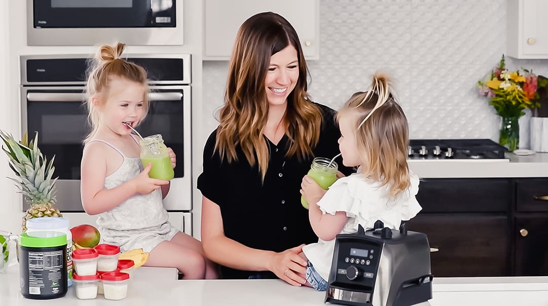 How to make the best green smoothie recipe! A simple green smoothie for beginners that is healthy, tastes amazing, and it's made with 4 ingredients in less than 5 minutes! Plus my tips on meal prepping smoothies! #greensmoothie #smoothie #smoothierecipe #greensmoothierecipe #kidfriendly front view of a mom and her two kids sitting on the kitchen counter drinking green smoothies